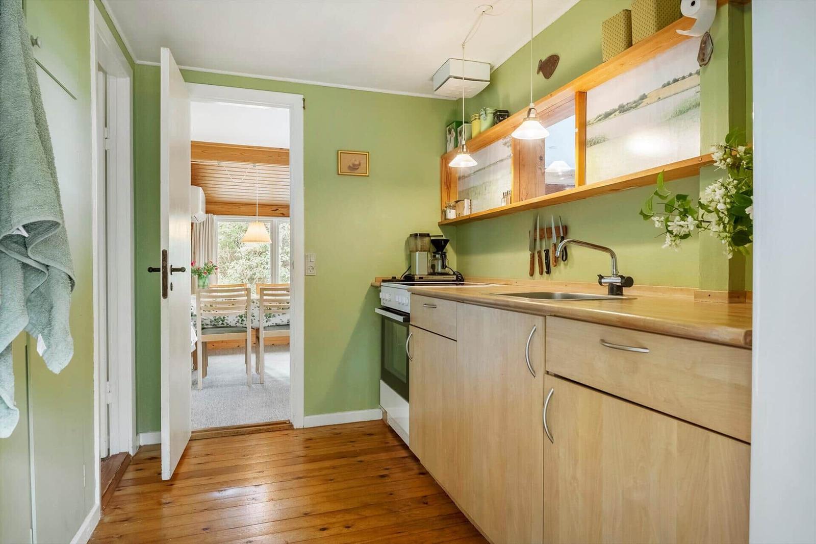 Küche mit Holzschrank, Arbeitsplatte und Spüle. Tür führt in Esszimmer mit Tisch und Stühlen.