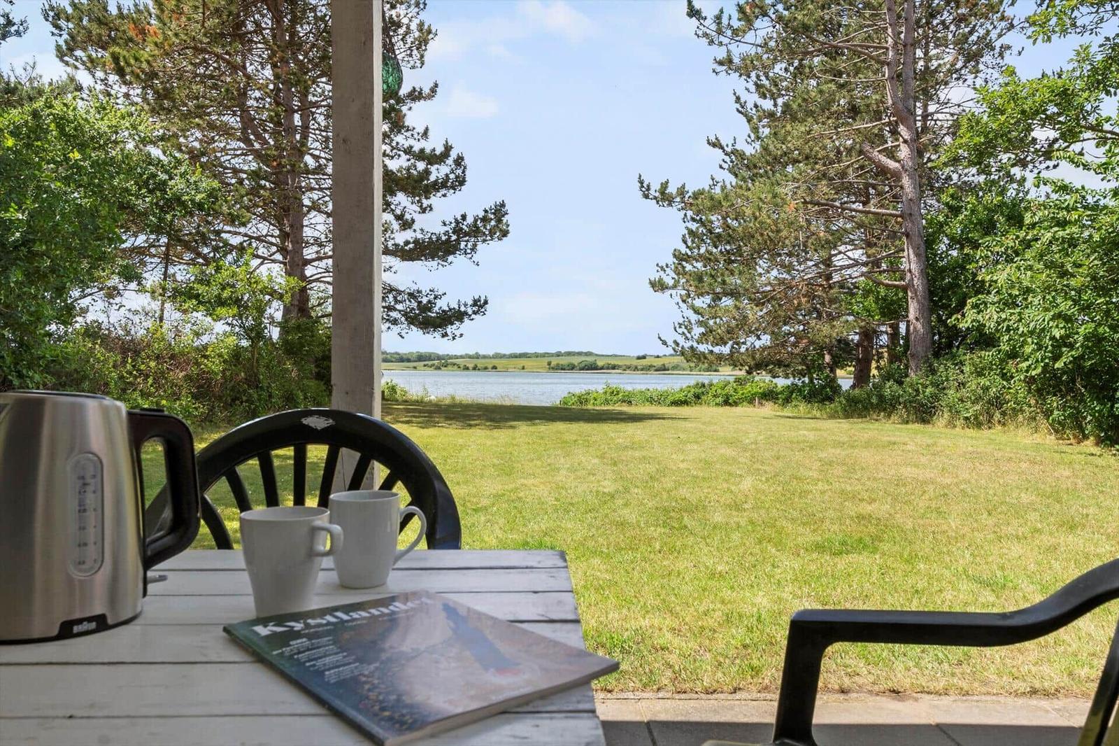 Terrasse mit Tisch, Stühlen und Blick auf Grasland und Wasser.