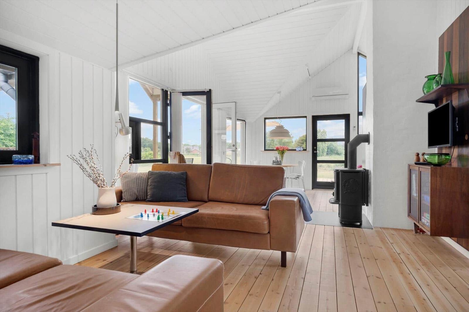 Wohnraum mit braunem Sofa, Holzboden und großen Fenstern. Kamin und TV-Möbel sind sichtbar.
