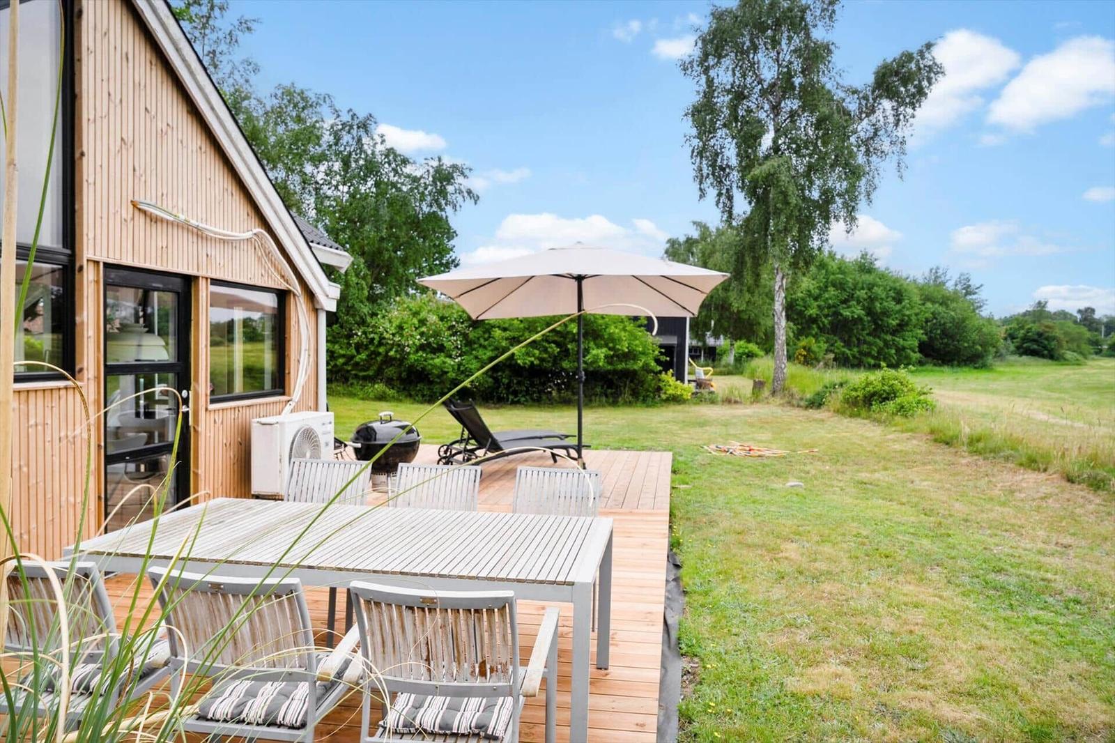 Terrasse mit Tisch, Stühlen und Sonnenschirm neben Holzhäuser. Grasfläche und Bäume im Hintergrund.