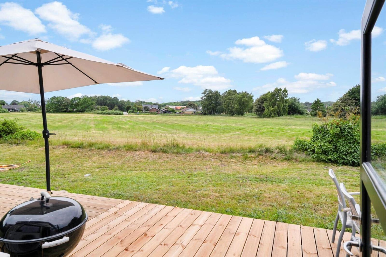 Terrasse mit Sonnenschirm und Grill, Blick auf grünes Feld und Bäume.