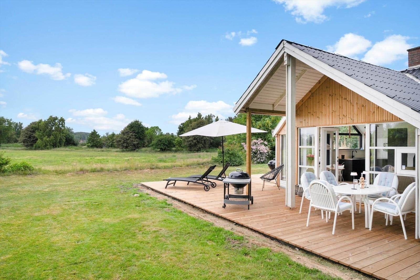 Terrasse mit Tisch, Stühlen, Grill und Liegen. Grünfläche und Wald im Hintergrund.