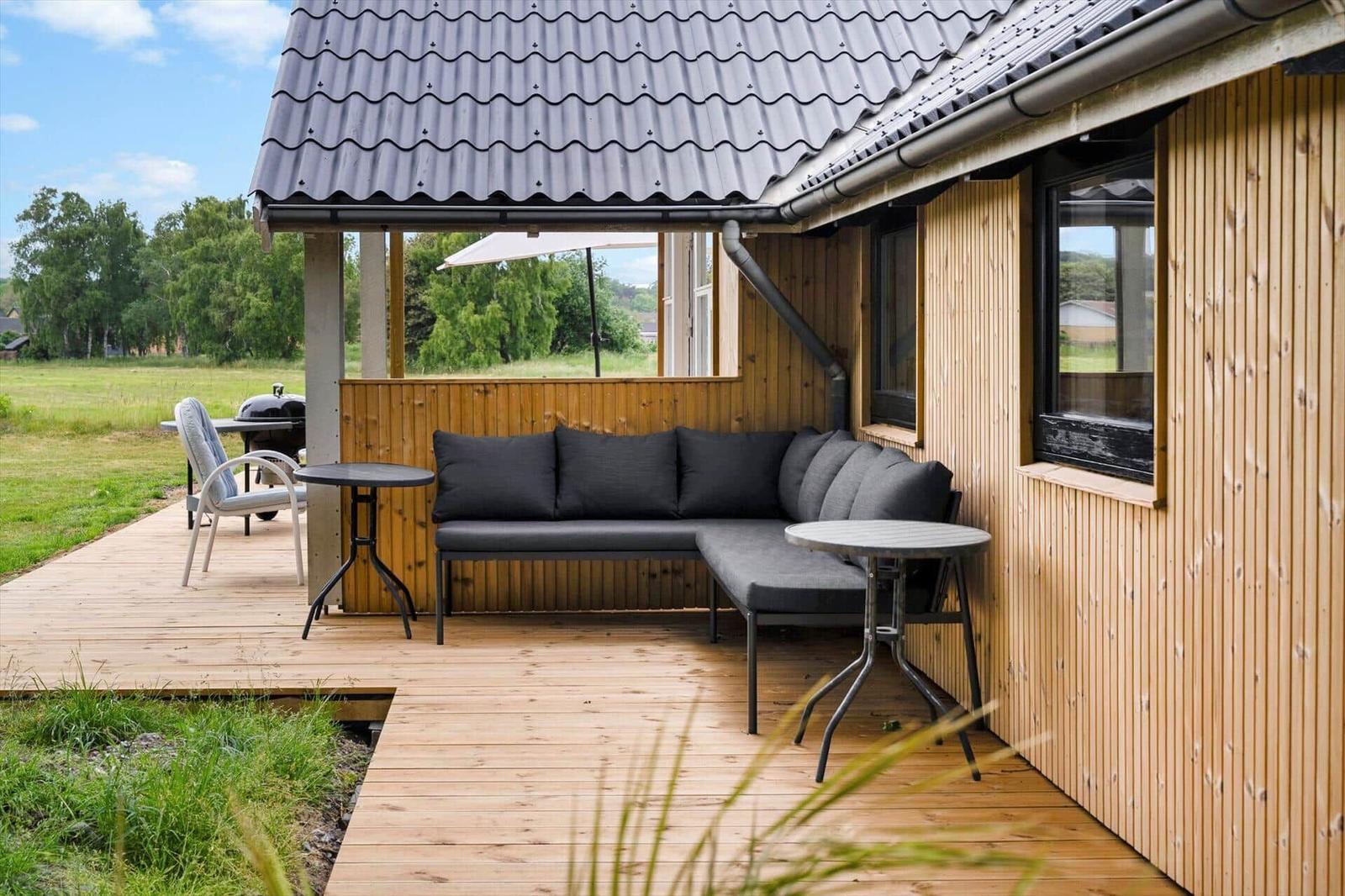 Terrasse mit Sofa, Tischen und Grill unter Dach. Holzwand und Grün im Hintergrund.