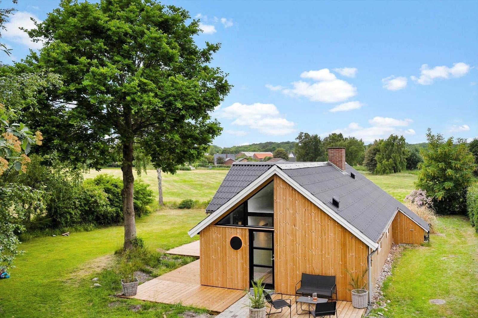 Holzhütte mit Terrasse und Garten. Blick auf Felder und Bäume unter blauem Himmel.