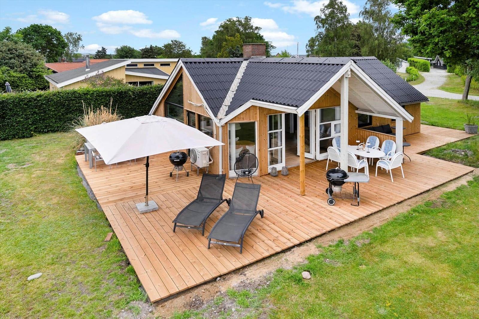 Holzhaus mit Terrasse, Sonnenliegen, Grill und Sitzgruppe unter einem Vordach.