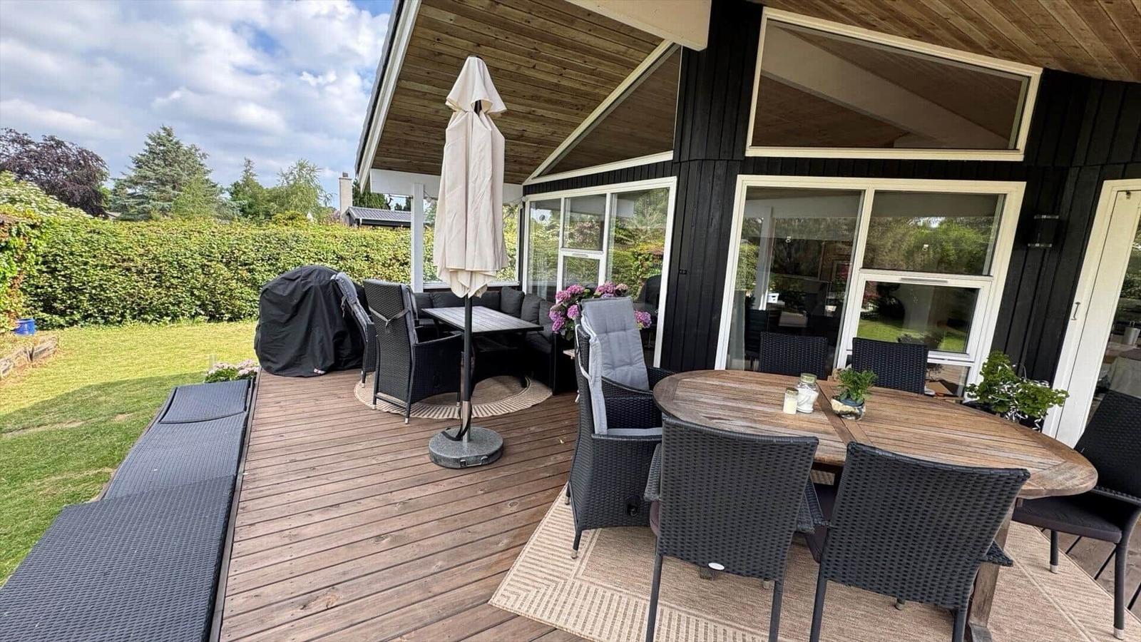 Terrasse mit Tisch, Stühlen und Sonnenschirm. Grünfläche und Hecke im Hintergrund.