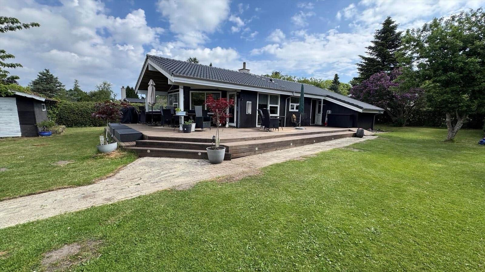 Moderne Villa mit großem Holzdeck, Garten und Sitzgelegenheiten unter freiem Himmel.