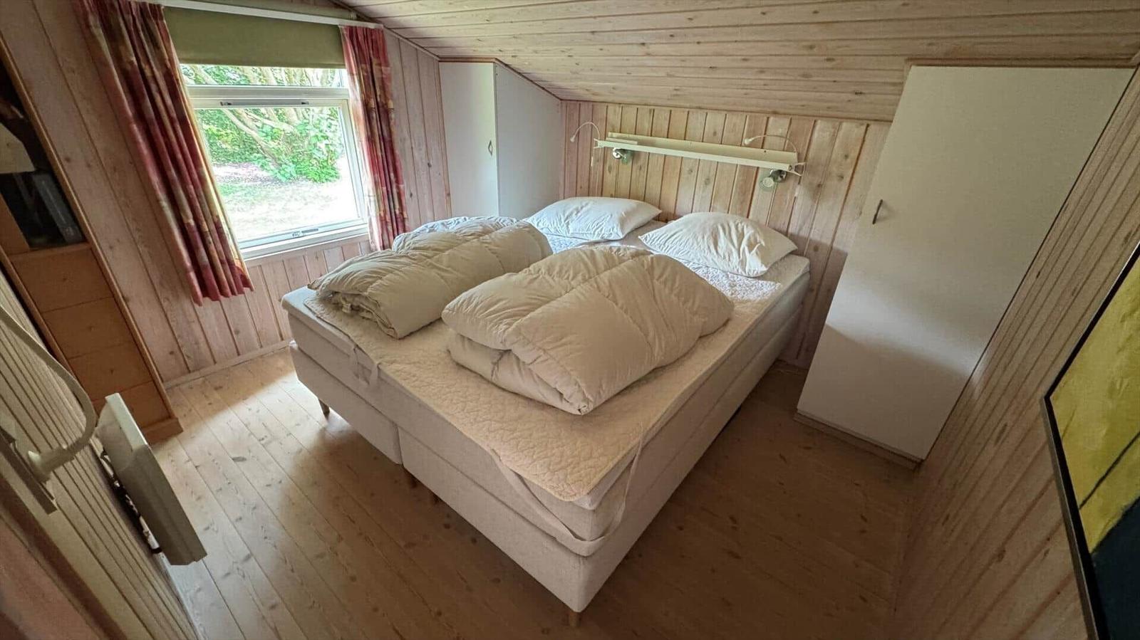 Schlafzimmer mit Doppelbett, Holzwand und Fenster mit Blick auf Grün.