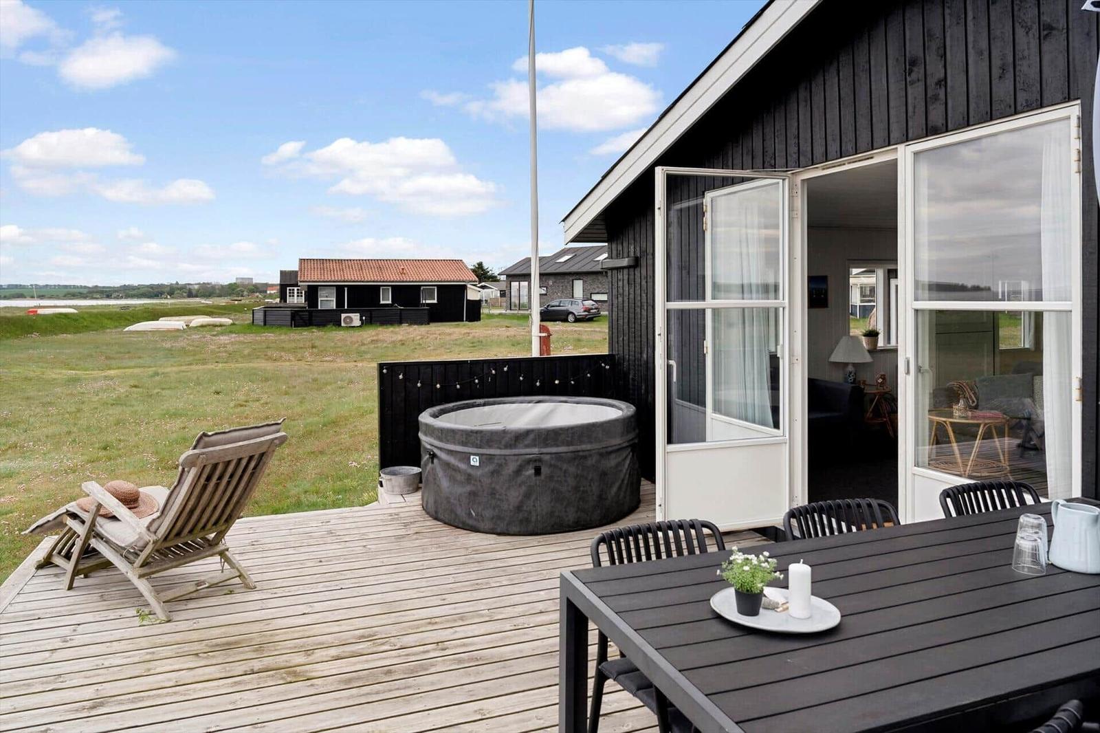 Terrasse mit Hot Tub, Esstisch und Liegestuhl. Blick auf grünes Feld und Haus.