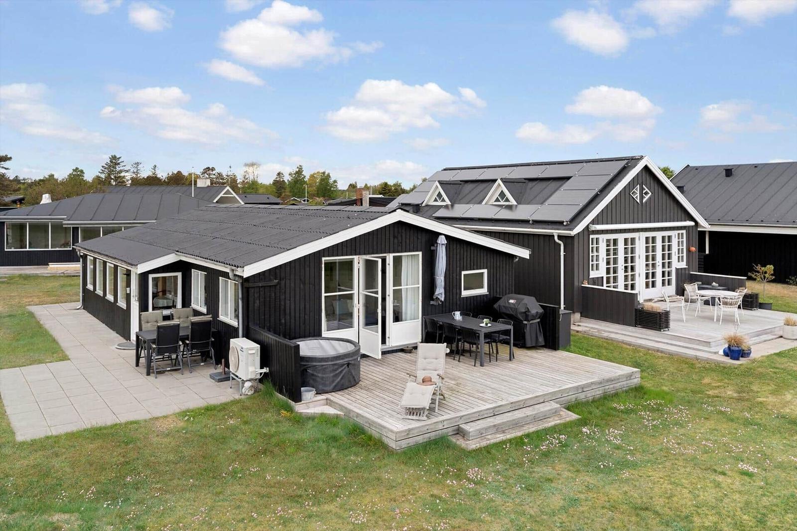 Schwarze Holzhütte mit Terrasse, Grill und Whirlpool. Grünflächen und klarer Himmel.