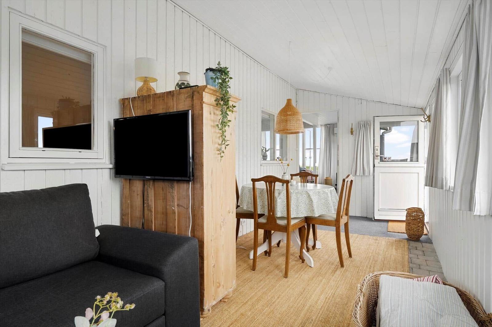Wohn- und Essbereich mit Sofa, TV, Tisch und Stühlen. Weiße Wände und Holzdecke.