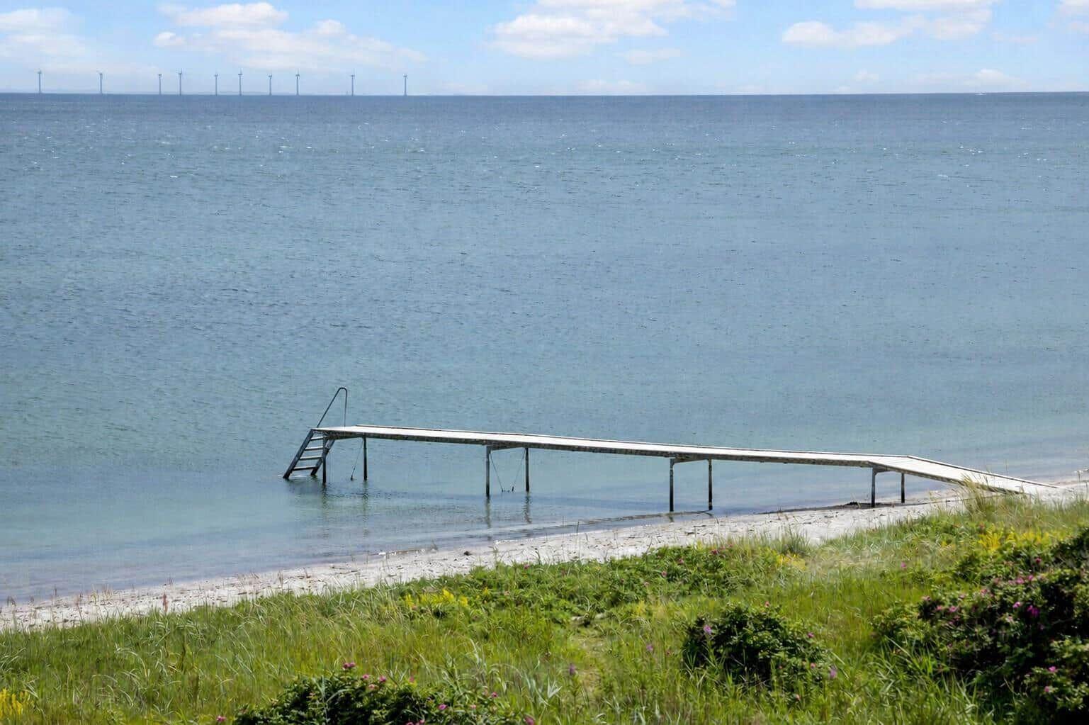 Ein hölzerner Steg führt zum Wasser. Im Hintergrund sind Windräder auf dem Meer sichtbar.