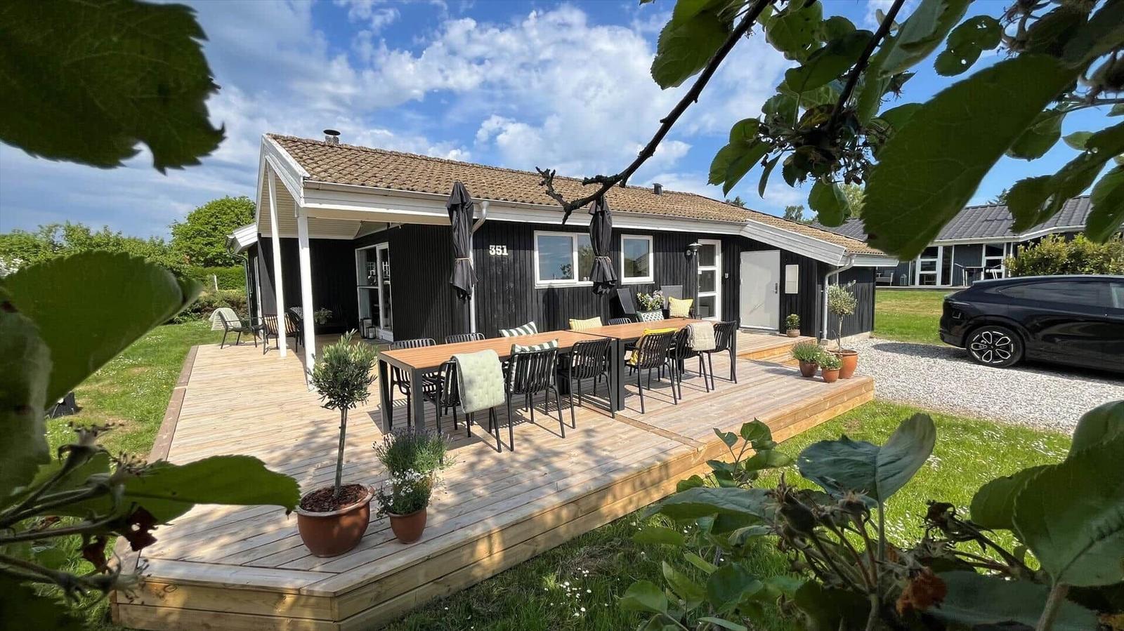Haus mit Holzterrasse, Tisch und Stühlen, Pflanzen und Grillplatz im Freien.