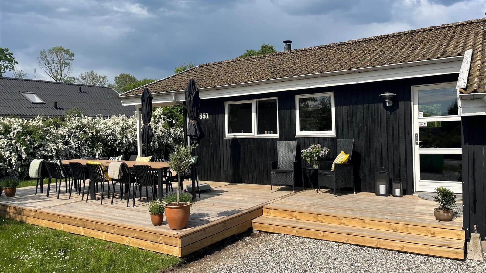 Schwarze Holzhütte mit Holzterrasse, Gartenmöbeln und Pflanzen. Hinterhof mit Beete und Himmel.