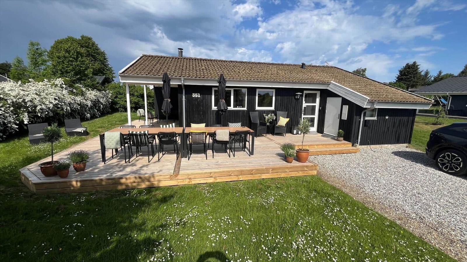 Schwarzes Haus mit Holzterrasse, Garten und Grillplatz. Ein Auto steht auf Kies.
