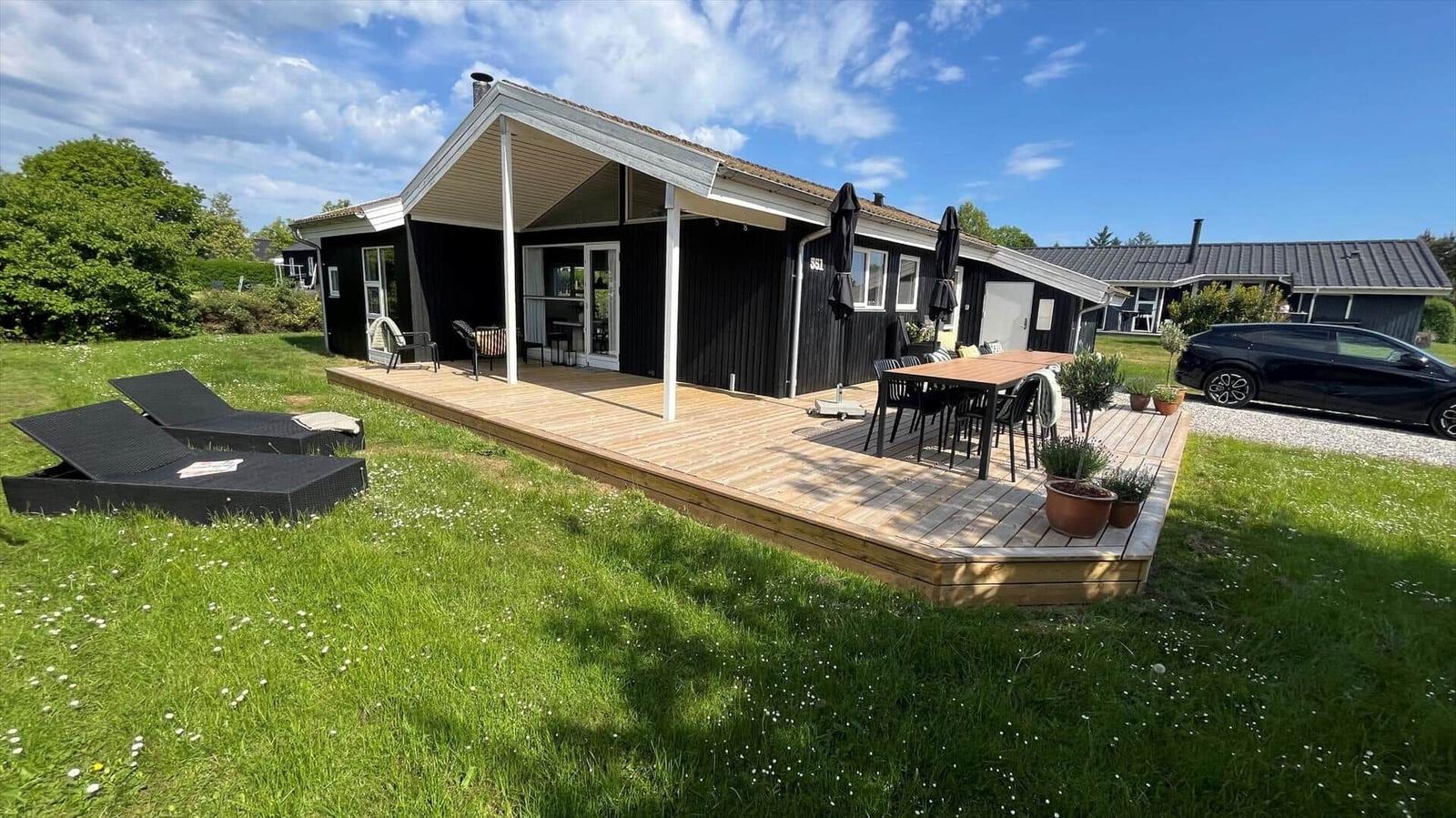Schwarzes Haus mit Holzterrasse, Liegen und Tisch. Grasfläche mit Blumen.