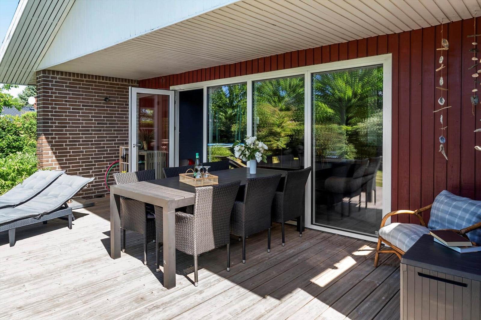 Terrasse mit Tisch, Stühlen und Liegen. Glasfront führt ins Haus. Holzdielen und rote Fassade.