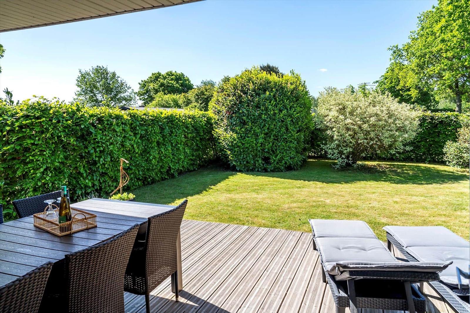 Terrasse mit Tisch, Stühlen und Liegen. Grünfläche und Hecke im Hintergrund.