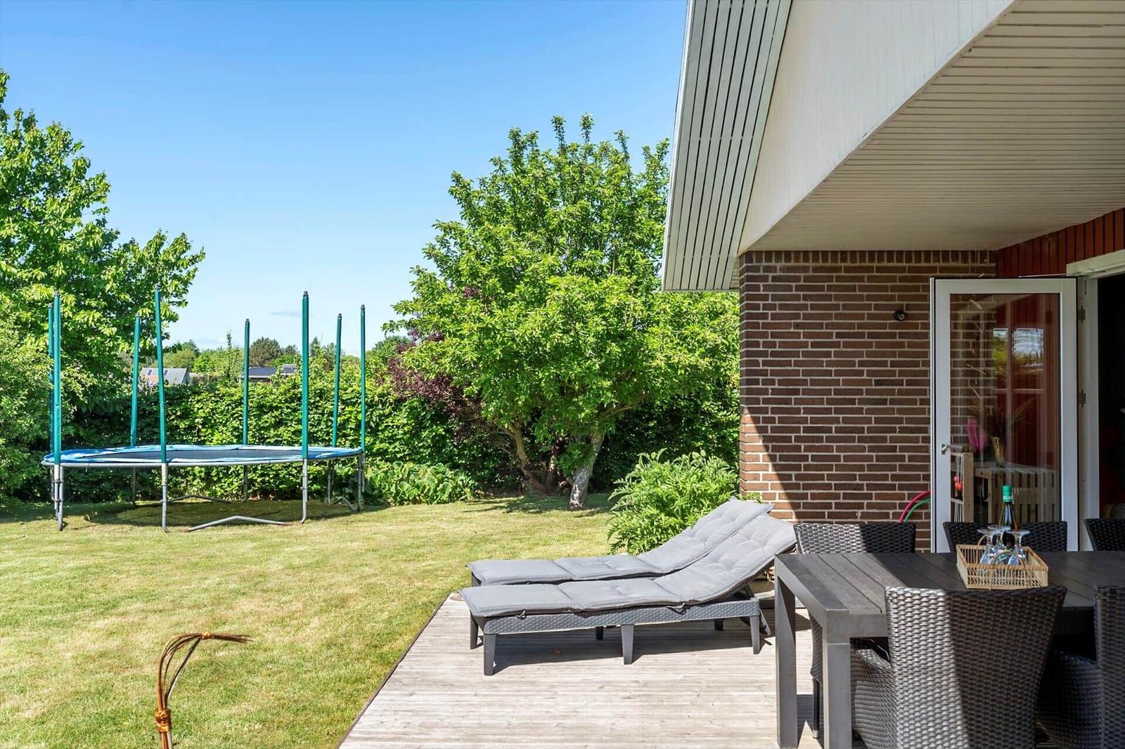 Terrasse mit Liegestühlen, Tisch und Trampolin im Garten. Haus mit Backsteinwand und Terrassendach.