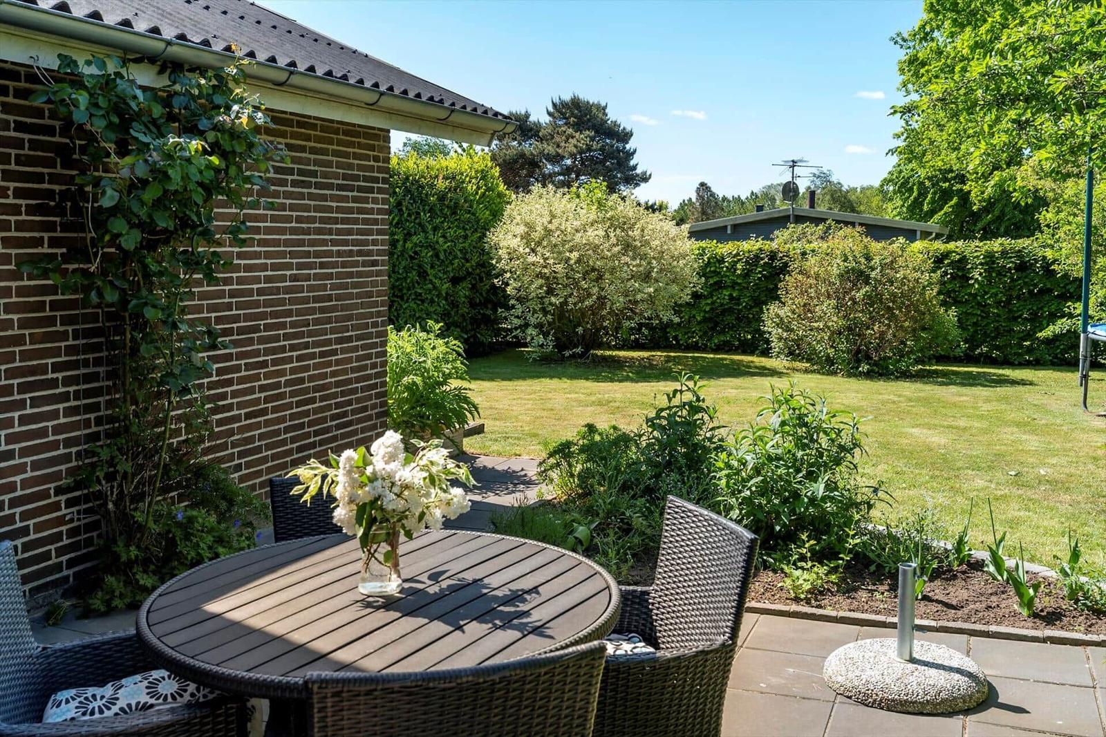 Runder Tisch mit Stühlen und Blumen auf Terrasse mit Garten und Hecke.