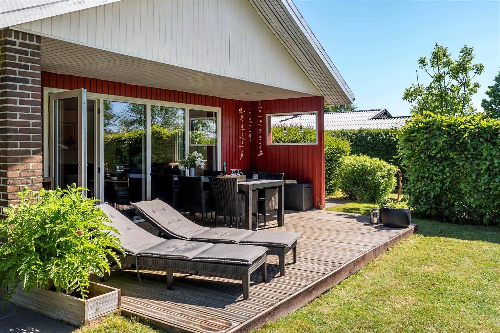 Terrasse mit Liegestühlen und Tisch. Roter Holzaufbau, Glasfront, grüne Pflanzen und Rasen.