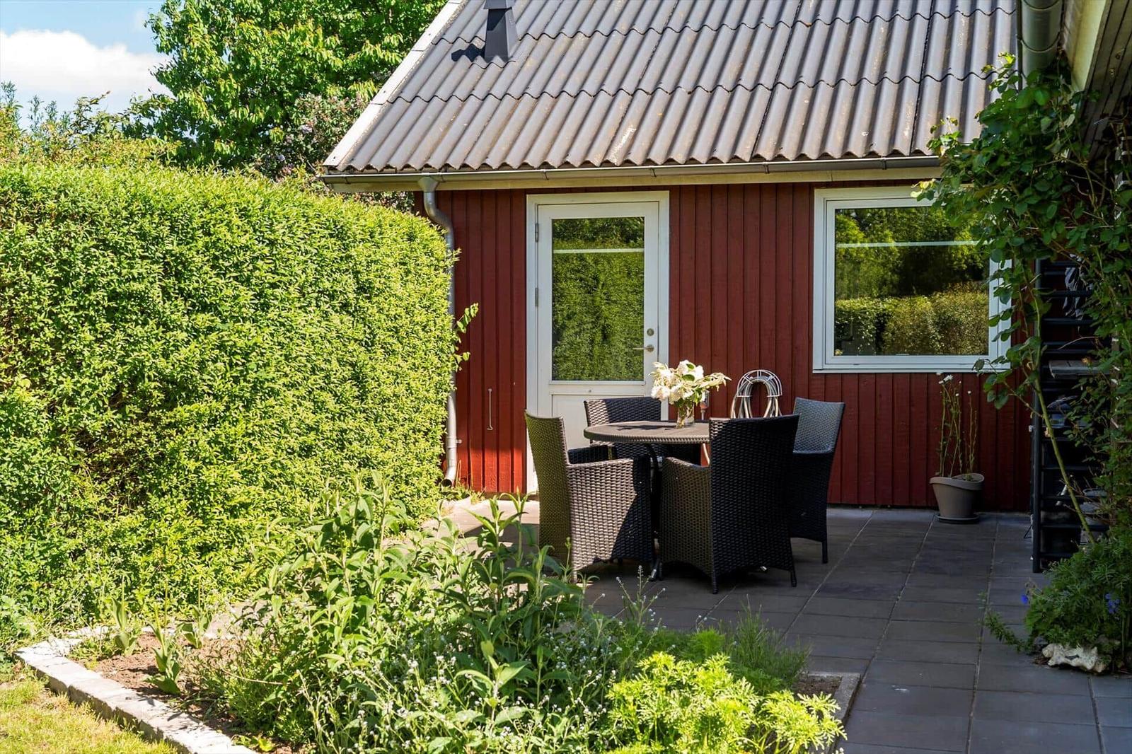 Roter Holzhaus mit Terrasse, Tisch und Stühlen, umgeben von grüner Hecke und Blumenbeeten.