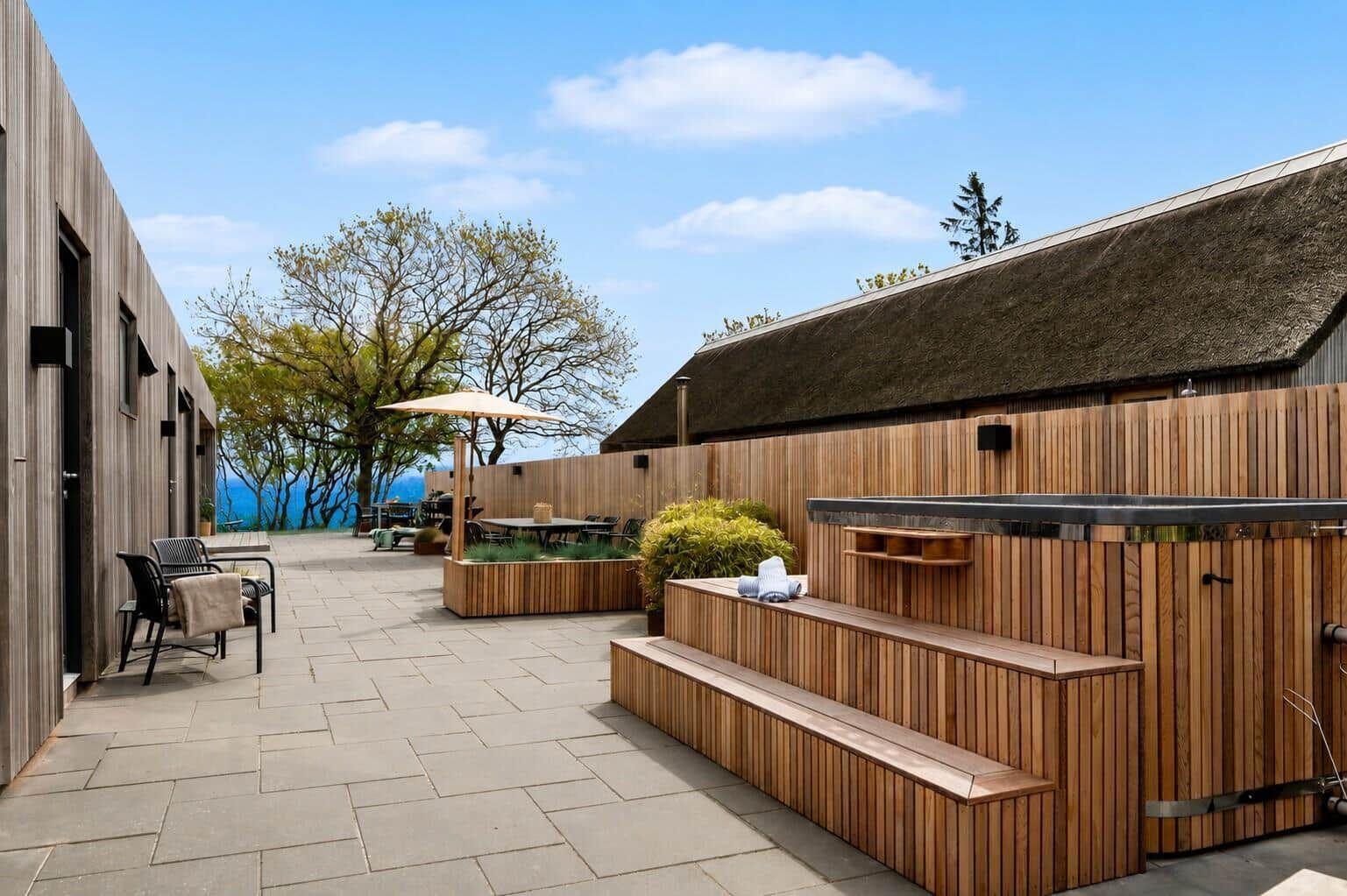 Außenbereich mit Holzsauna, Sitzbank und Terrasse. Blick auf Bäume und Himmel.