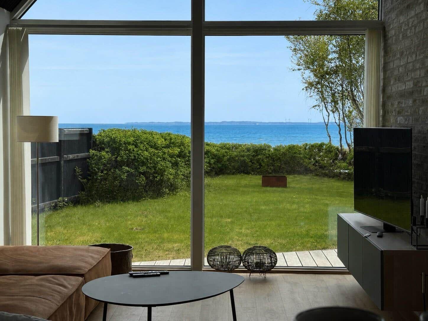 Wohnzimmer mit Blick auf Grünfläche und Meer. Große Fenster, Sofa, Couchtisch, Fernseher.