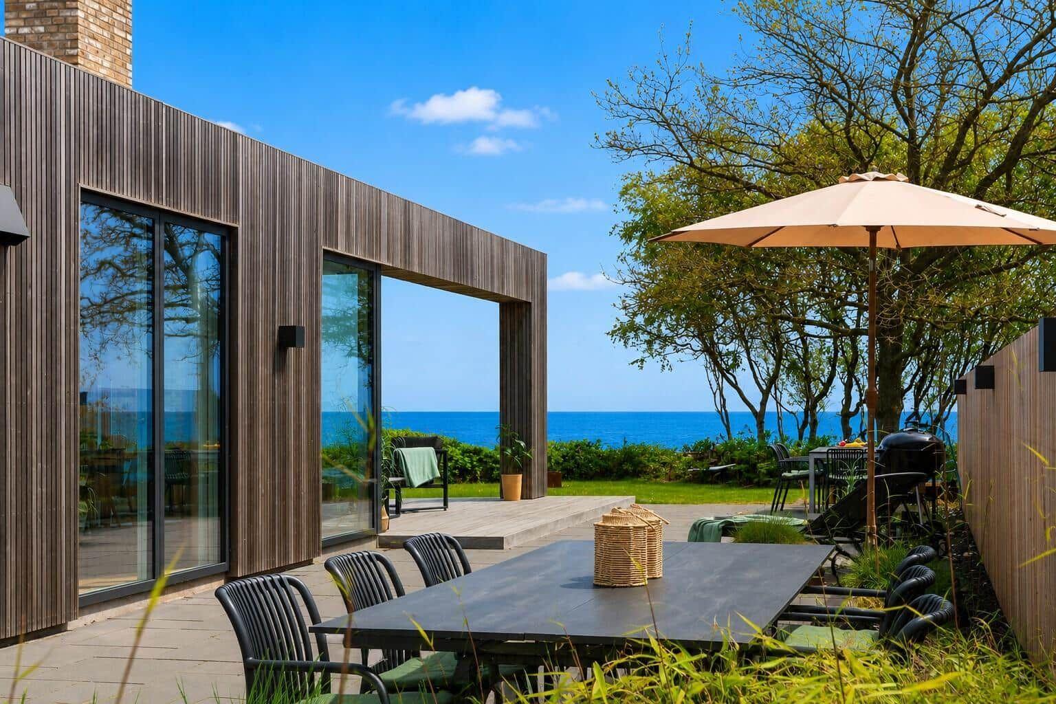 Terrasse mit Tisch, Stühlen und Sonnenschirm. Blick auf Meer und Grünflächen.