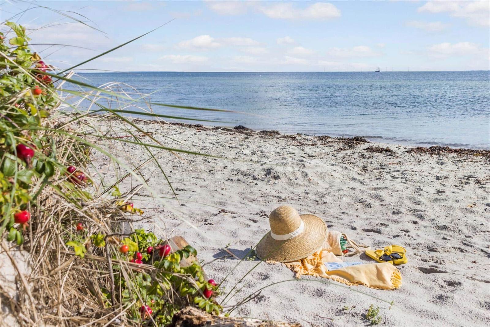 Strand mit Strohhut, Handtuch und Sonnenbrille. Blick auf das Meer mit Segelyacht im Hintergrund.