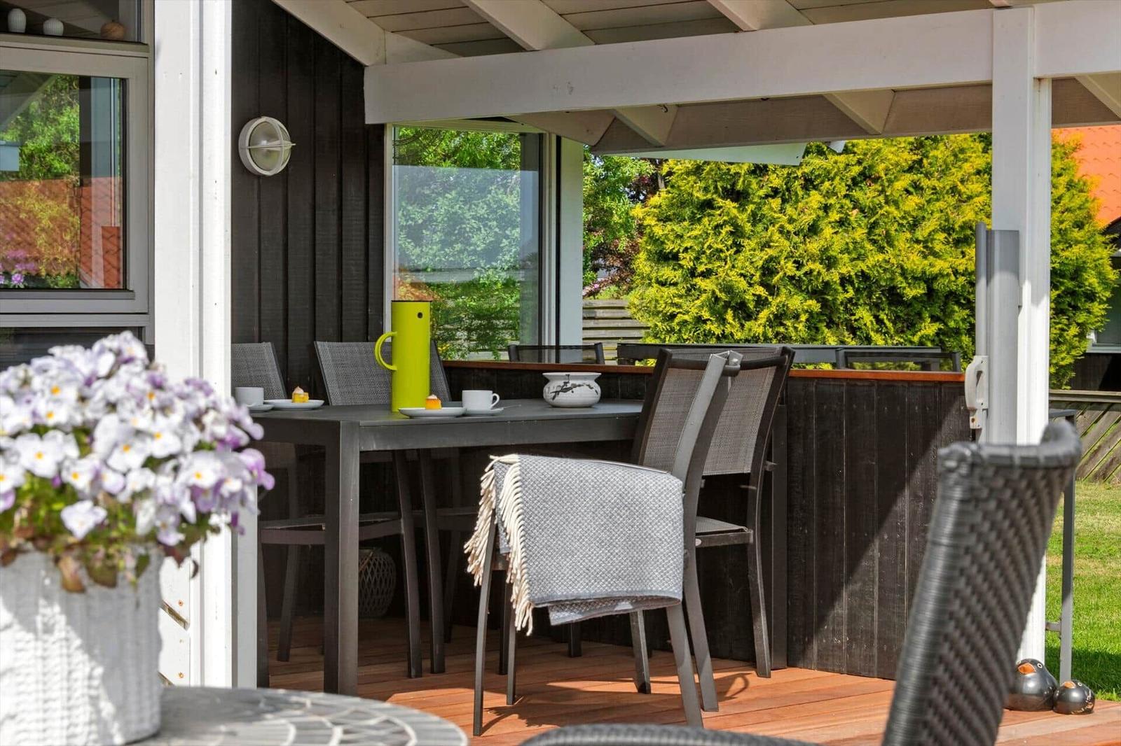 Terrasse mit Tisch, Stühlen und Blumen. Hintergrund: grüne Hecke und Holzwand.