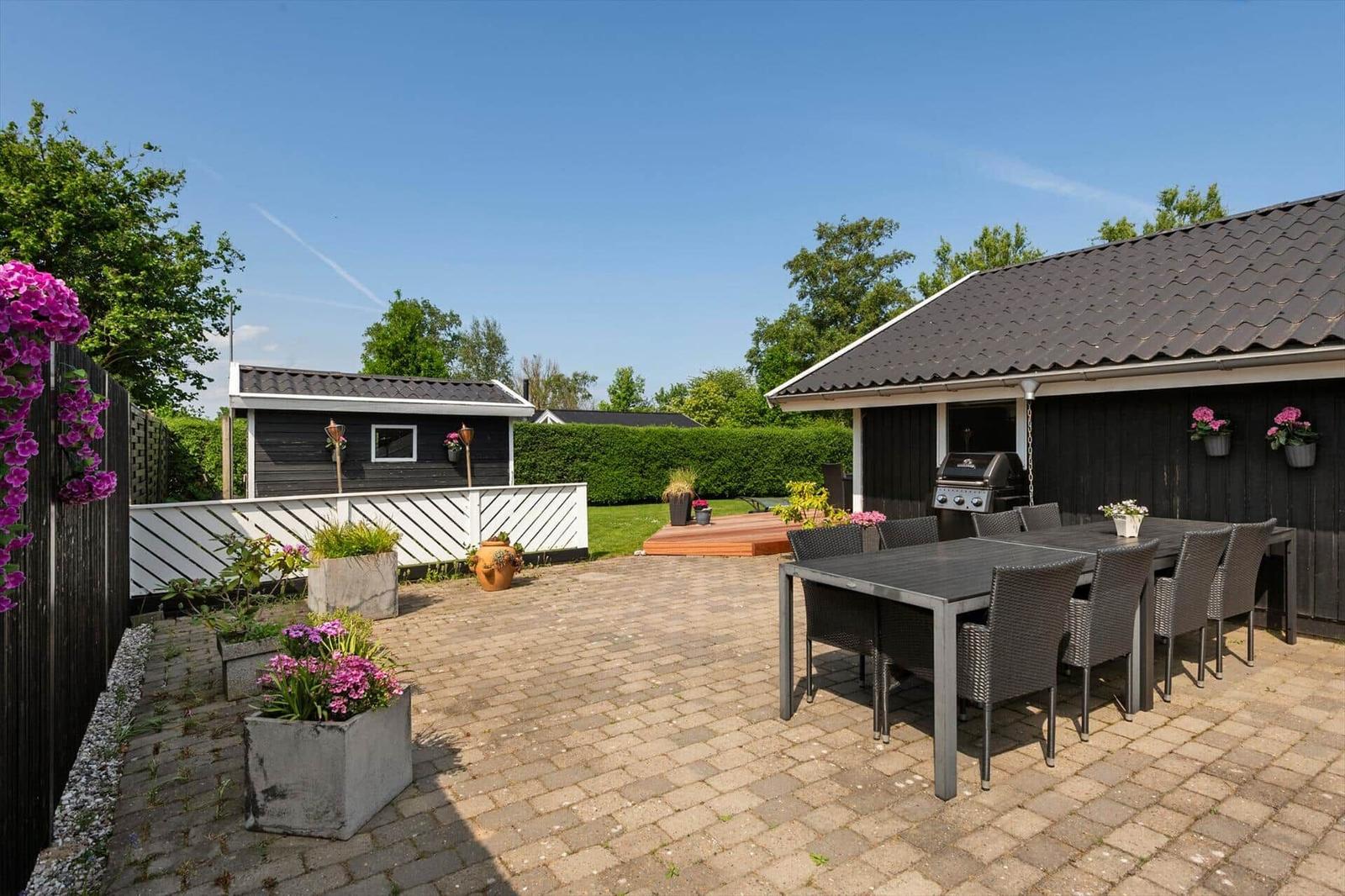Terrasse mit Grill, Tisch und Stühlen. Umgeben von Blumen und Grünflächen.