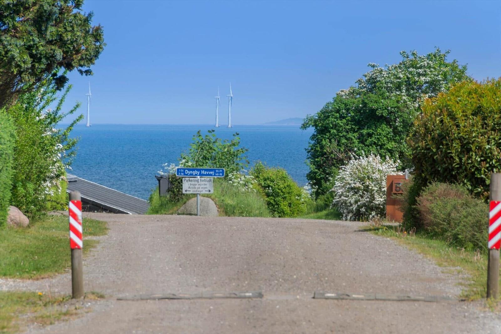 Einfahrt mit Blick auf das Meer und Windkraftanlagen. Straßenschild und Grünflächen sind sichtbar.