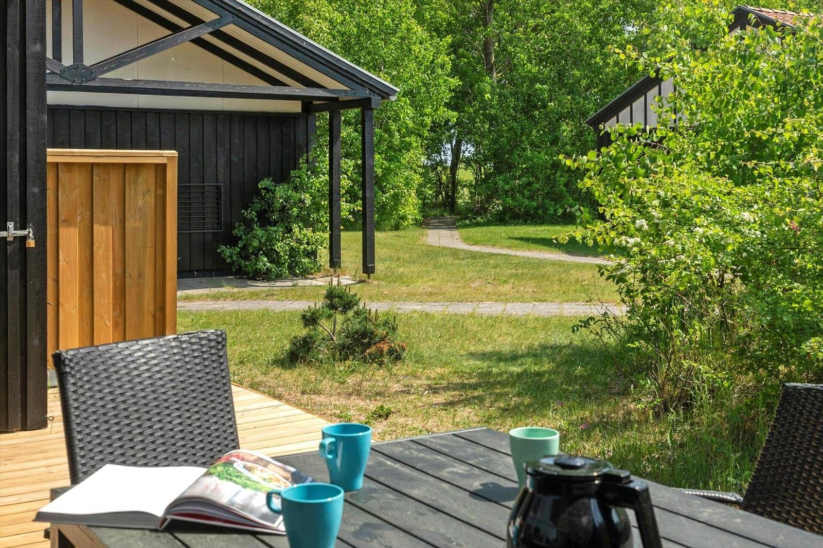 Terrasse mit Tisch, Stühlen und Kaffeekanne. Grünflächen und Bäume im Hintergrund.