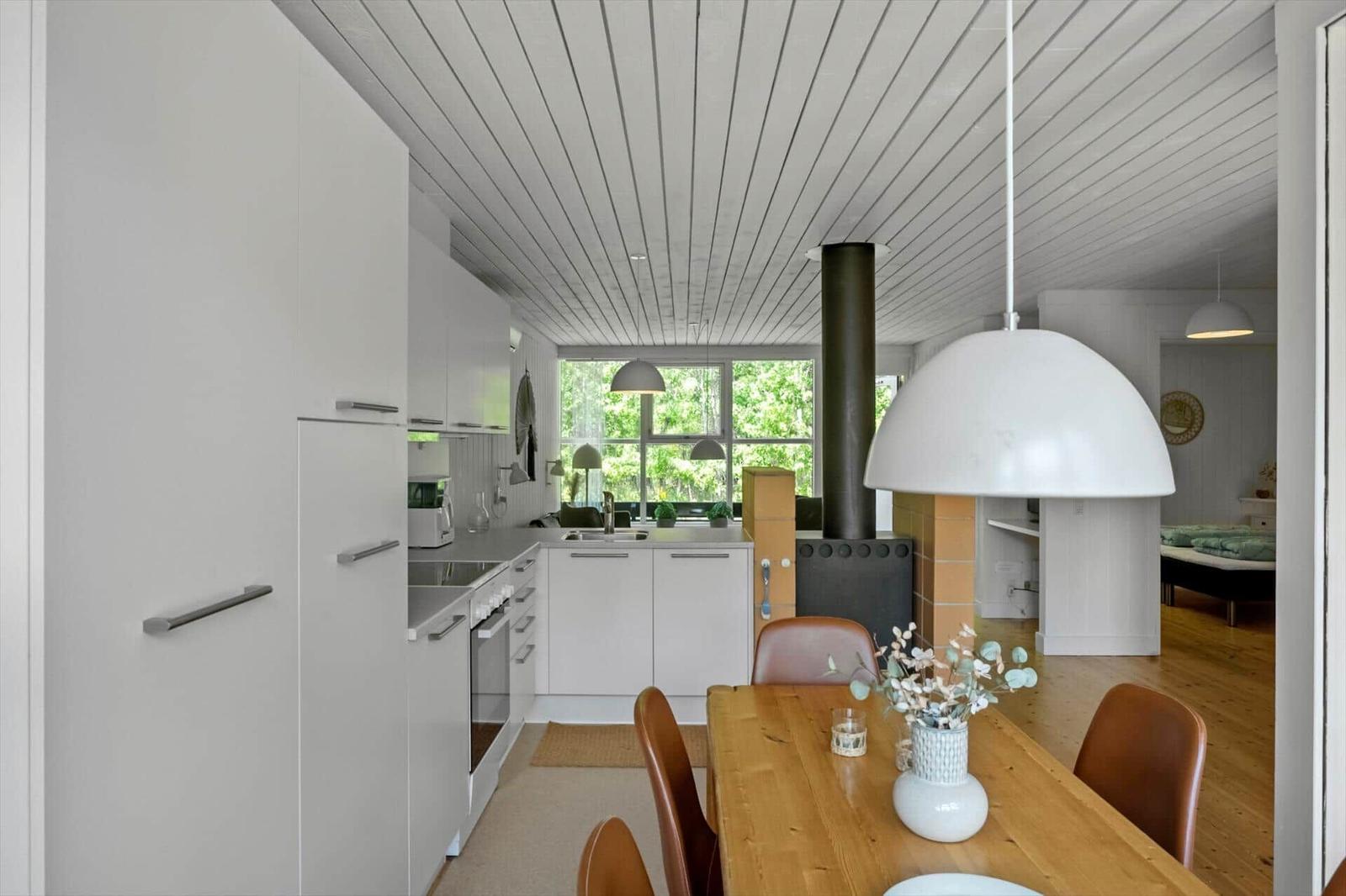 Küche mit Essbereich und Holzboden. Große Fenster und weiße Schränke. Zentraler Kamin mit Schornstein.