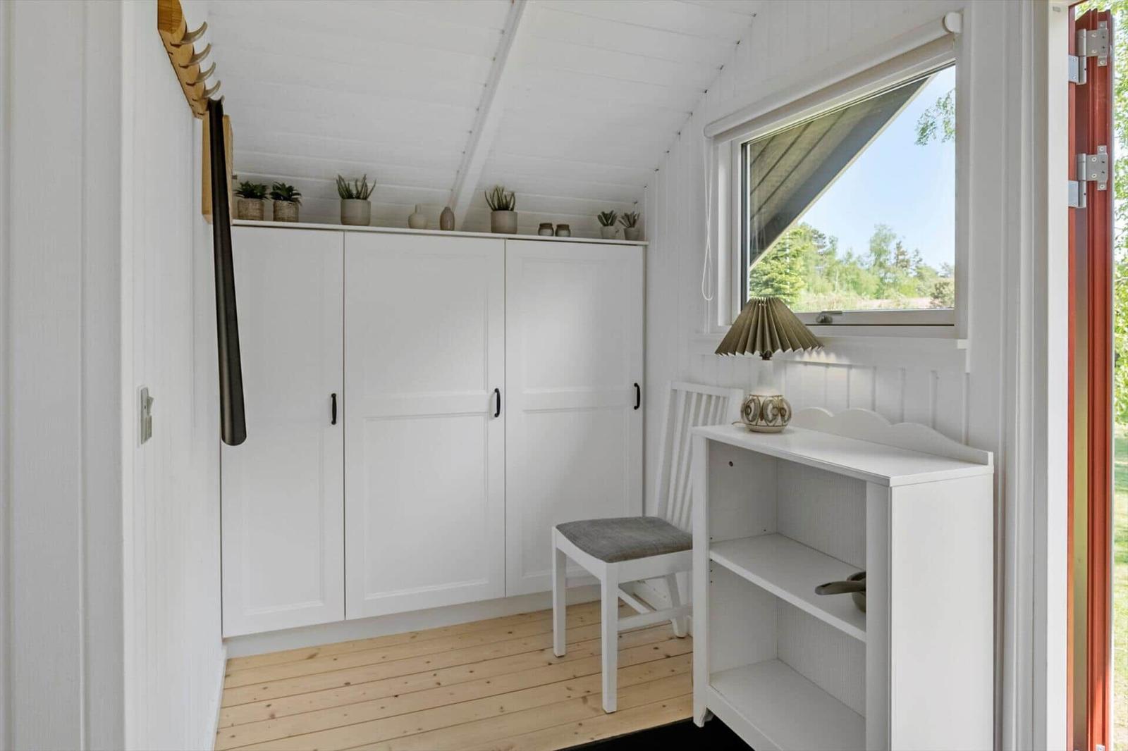 Room with white closet, chair, and shelf. Window with view of green.