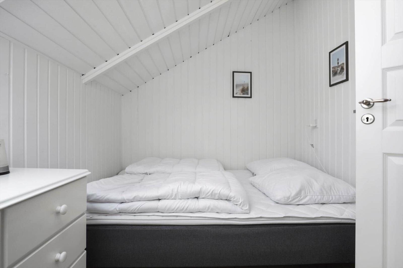 Bedroom with white bedding, wooden walls, and dresser. Door and pictures are visible.