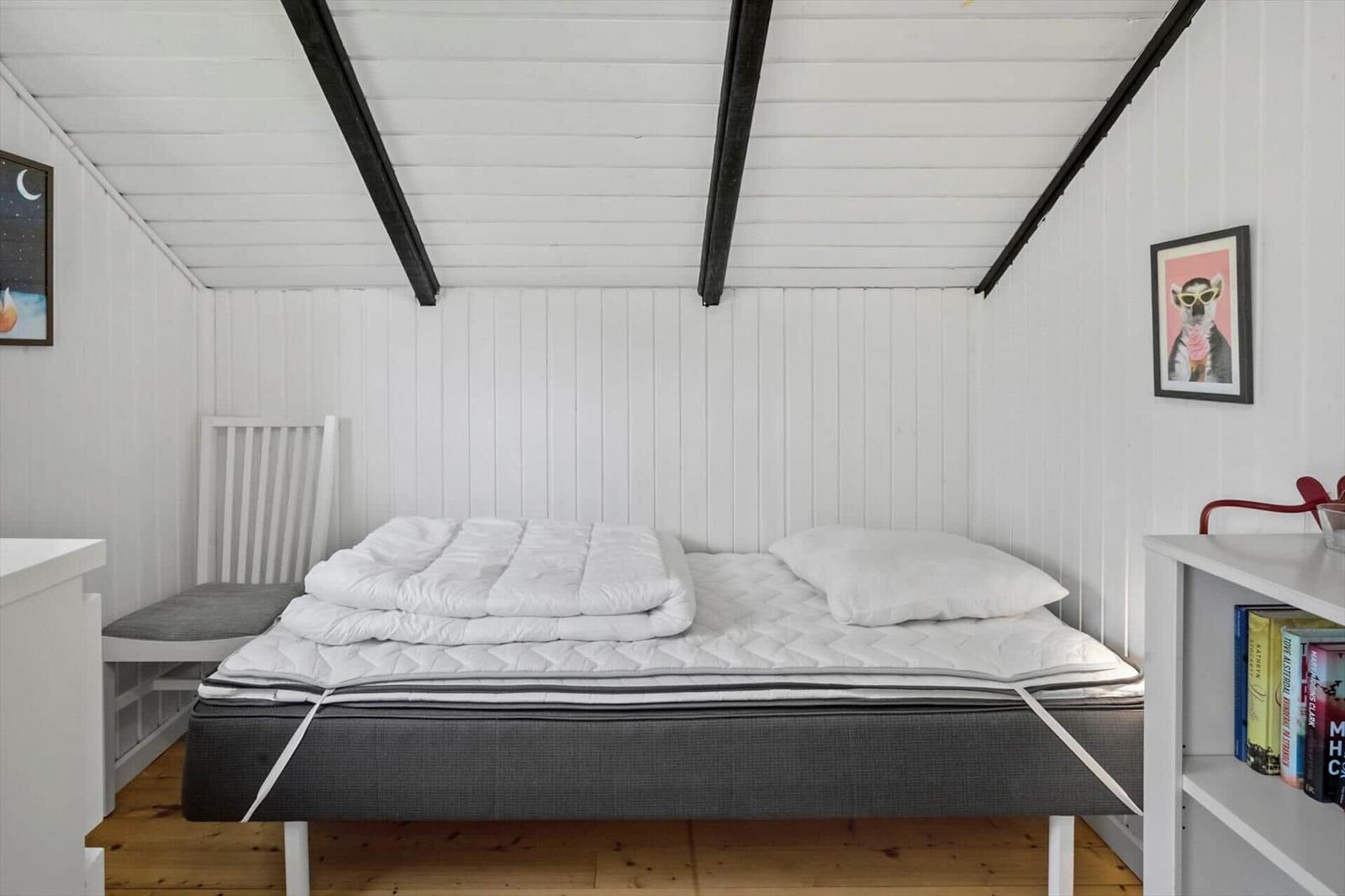 Bedroom with bed, chair, and shelf. White walls, wooden floor, and black beams.