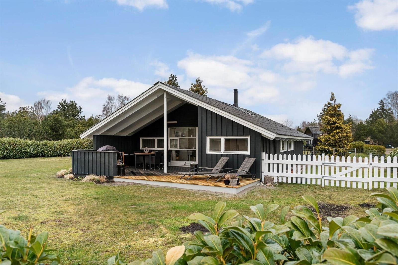 Ein Haus mit Holzterrasse, Liegestühlen und weißem Zaun auf grünem Grund.