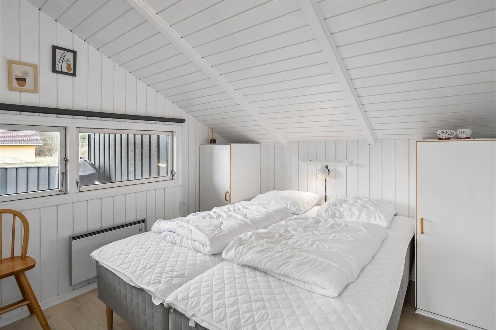 Schlafzimmer mit Doppelbett, weißen Wänden und Dachschrägen. Fenster, Schrank und Stuhl sichtbar.
