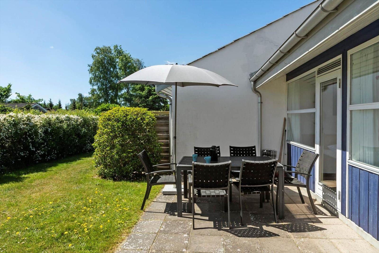 Terrasse mit Tisch, Stühlen und Sonnenschirm. Grasfläche und Hecke im Hintergrund.
