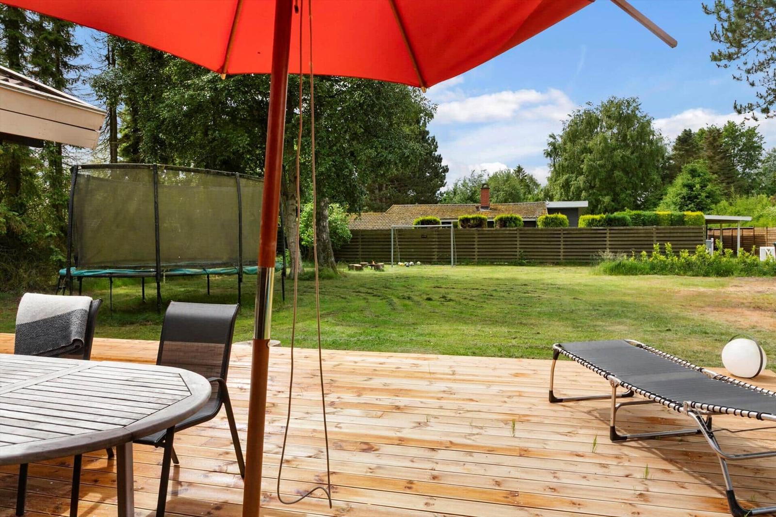 Holzterrasse mit Tisch, Stühlen und Liege unter rotem Sonnenschirm. Grasfläche und Trampolin im Hintergrund.