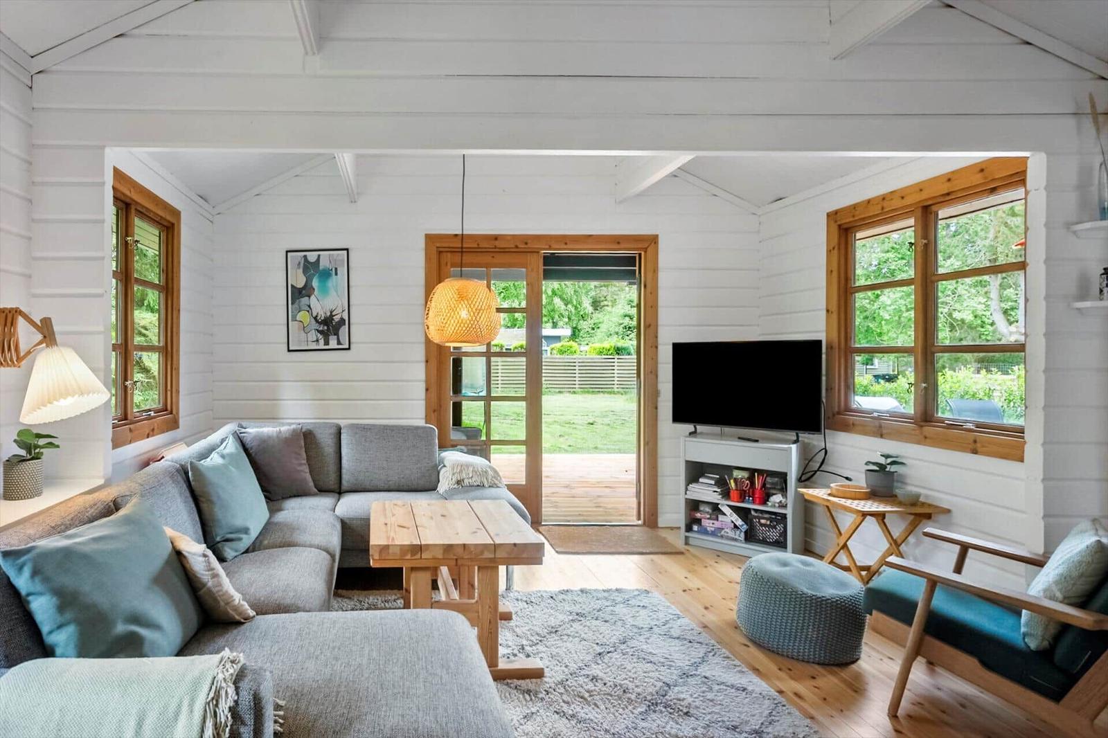 Wohnzimmer mit Sofa, Tisch und TV. Holzfenster und Tür zu Garten.