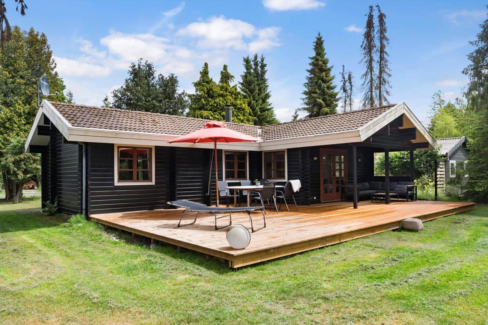 Schwarzes Holzhaus mit Terrasse, Sonnenschirm und Gartenmöbeln unter blauem Himmel.