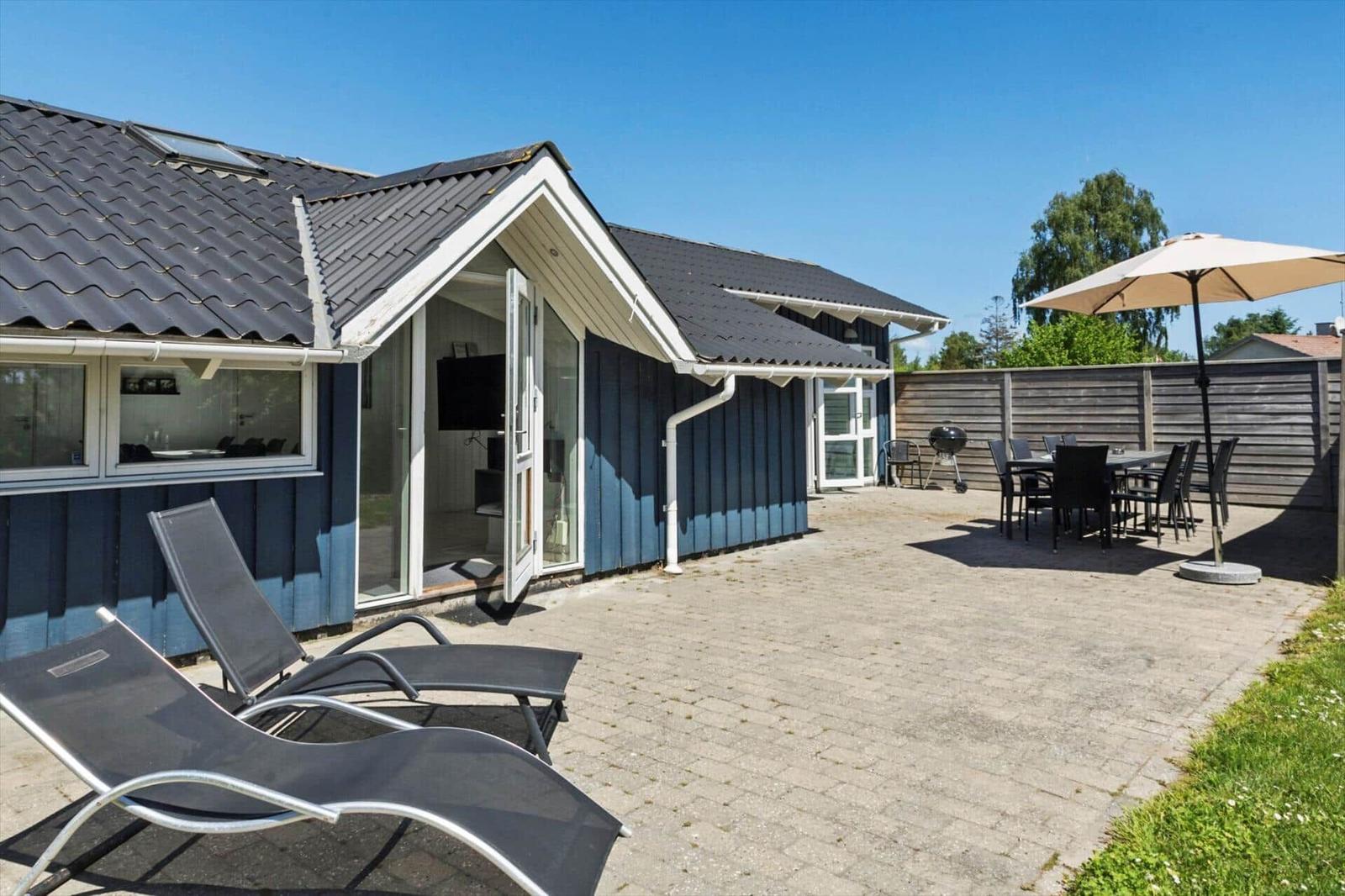 Blau gestrichene Ferienwohnung mit Terrasse, Liegestühlen, Grill und Tisch mit Sonnenschirm.