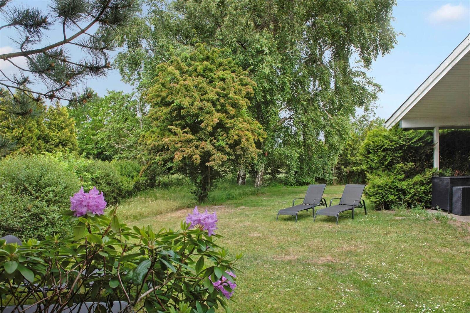 Garten mit Liegestühlen, Blumen und Bäumen. Teil eines Hauses mit Überdachung.