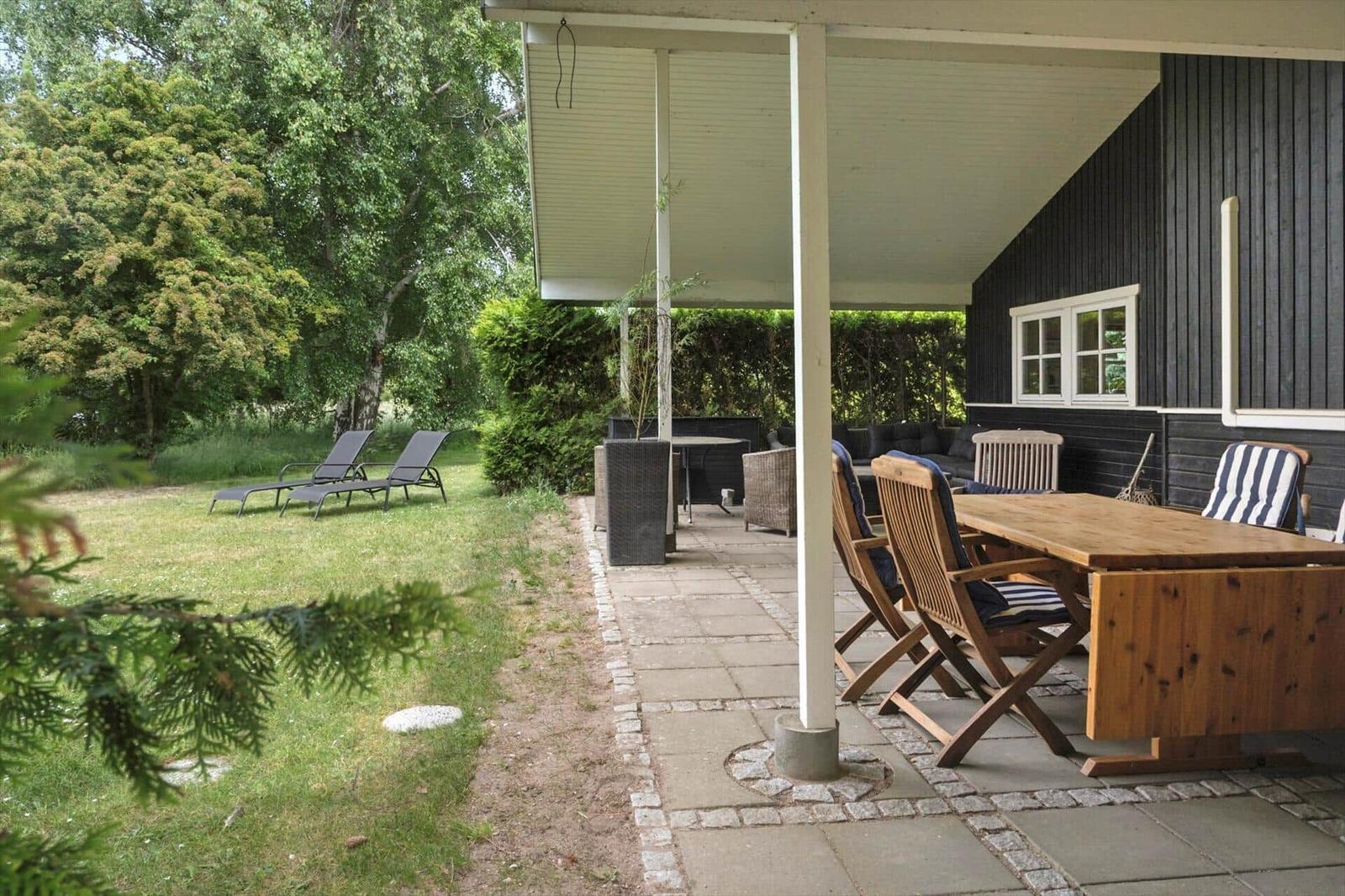 Terrasse mit Holztisch, Stühlen und Liegestühlen im Garten. Schwarze Hauswand mit Fenster.