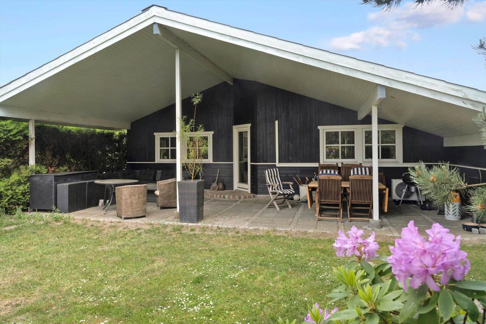 Haus mit überdachter Terrasse, Gartenmöbeln und Blumen im Vordergrund.