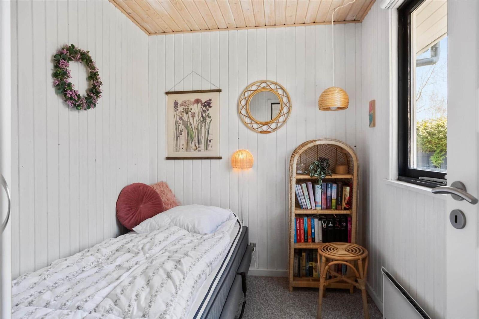 Ein Schlafzimmer mit Bett, Bücherregal und Fenster. Wände sind weiß verkleidet.