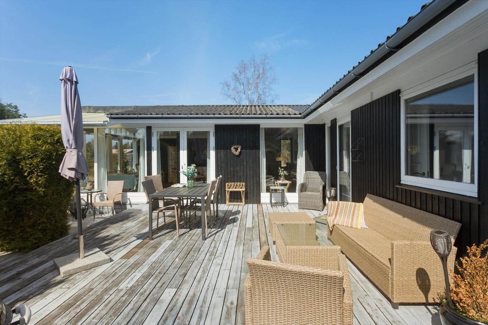 Terrasse mit Sitzgruppe, Tisch und Sonnenschirm. Haus mit Glasfront und dunkler Fassade.